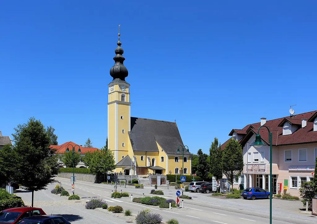 Südansicht des Dorfplatzes mit der katholischen Pfarrkirche hl. Petrus der oberösterreichischen Gemeinde Moosbach. Ein gotischer Sakralbau mit einer vorwiegend neogotischer Einrichtung aus den Jahren 1902 bis 1906. Der Westturm wurde 1766 erhöht und eine barocke Zwiebelhaube aufgesetzt.