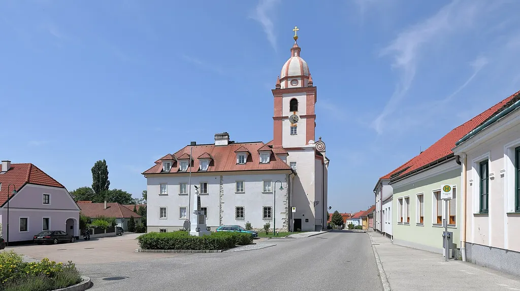 Ostansicht des Ortszentrums von Maria Roggendorf, ein Ortsteil der niederösterreichischen Marktgemeinde Wullersdorf, mit der ortsbildbeherrschenden Wallfahrts- und Pfarrkirche Mariä Geburt, dem angebauten Pfarrhof, der heute als Benediktinerkloster St. Josef genutzt wird, und davor das Kriegerdenkmal.