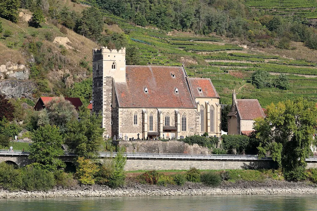 Südansicht (vom rechten Donauufer) der Filialkirche hl. Michael und des Karners (um 1395 erbaute) in St. Michael, ein Ortsteil der niederösterreichischen Marktgemeinde Weißenkirchen in der Wachau.Die ehemalige Wehrkirche wurde leicht erhöht am linken Donauufer errichtet und ist die Mutterpfarre der Wachau. Sie wurde an der Stelle einer kleinen keltischen Opferstätte errichtet und im Jahre 987 erstmals urkundlich erwähnt. Die Kirche in ihrer heutigen Gestalt ist ein spätgotischer, innen barockisierter Staffelhallenbau mit Chor und einem markanten Wehrturm aus dem ersten Viertel des 16. Jahrhunderts. Um 1500 bis 1530 wurde sie durch eine Wehranlage ergänzt bzw. diese verstärkt. Seit der Josephinischen Kirchenreform ist die Kirche eine Filiale von Wösendorf (1784).