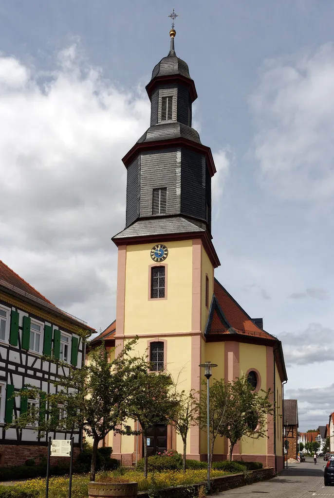 Evangelische Kirche in Dudenhofen, einem Stadtteil von Rodgau (Hessen). Das Foto zeigt die Frontalansicht mit Hauptportal und Kirchturm. Links ist das Rektor-Geißler-Haus, das ebenfalls unter Denkmalschutz steht.