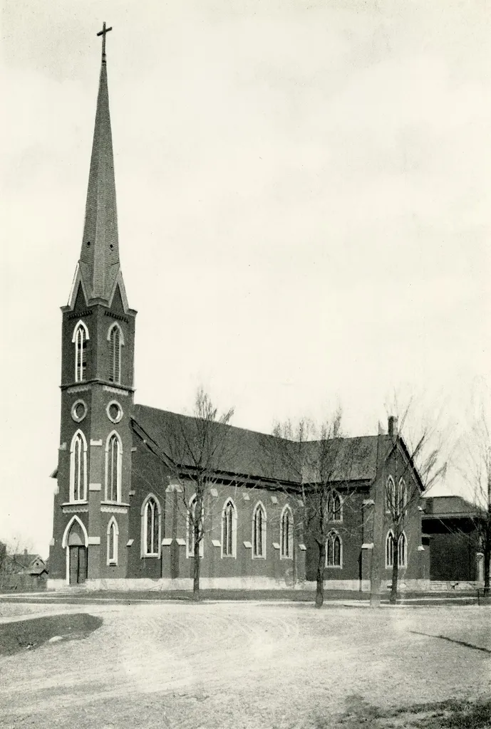 A winter photograph of the First Roman Catholic Church in Lansing, Michigan, from the perspective of the intersection of Madison and Chestnut Street. The wings on the east and west side were added in 1879, and the tower and spire of the church were completed in 1880. The parson's house can be seen behind the church, at the far right of the photo. The building was demolished in 1906.