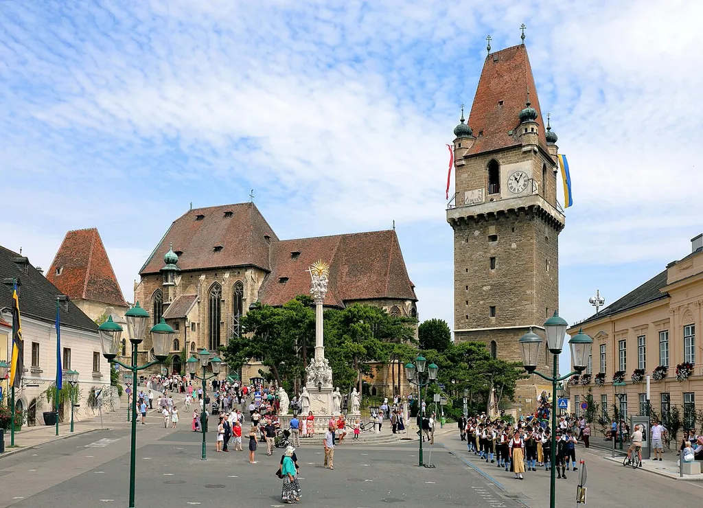 Der Marktplatz der niederösterreichischen Marktgemeinde Perchtoldsdorf. Links das sogenannte Regenharthaus, ein Bürgerhaus mit einer Kernsubstanz aus dem 15. Jahrhundert und hinter diesem die Martinskapelle bzw. der ehemalige Karner mit dem steilem Keildach. In der Bildmitte die Pfarrkirche hl. Augustinus und vor dieser die barocke Dreifaltigkeitssäule. Rechts der Wehrturm, ein von 1450 bis 1521 erbauter fünfgeschoßiger Turm, und ganz rechts das Gemeindeamt, ein ehemaliger späthistoristischer Meierhof aus dem Jahr 1838.Die Aufnahme wurde am Perchtoldsdorfer Fronleichnamstag gemacht, der gegenüber dem „regulären“ Fronleichnamstag um 10 Tage später gefeiert wird.