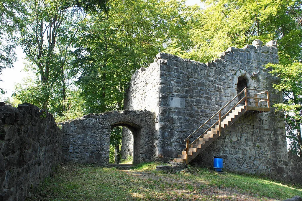 Innenhof der Ruine Ramsenburg, Blick Richtung Eingang (links) und hölzerner Treppe, die zum mit einem Gitter verschlossenen Eingang der Turm-Ruine (rechts) führt, in der Bildmitte eine steinerne Inschrift-Tafel in der Turm-Mauer