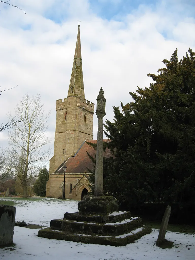 Stone cross in Holy Trinity parish churchyard, Belbroughton, Worcestershire, seen from the southeast, with the church steeple in the background, in snow. The cross is late Medieval, and was restored early in the 20th century as a monument to Major-General Sir Edward Woodgate, who was killed in 1900 in the Second South African War.