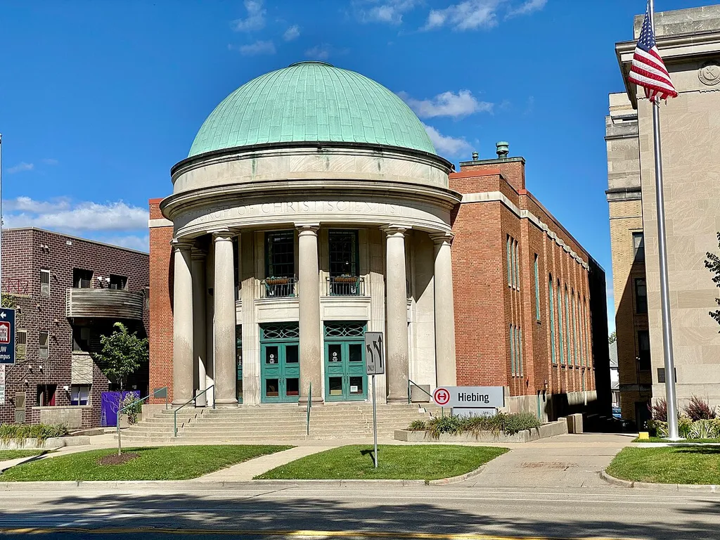 Built in 1929, this Classical Revival / Georgian Revival-style building at 315 Wisconsin Avenue in Madison, Wisconsin, was designed by Frank M. Riley to house the congregation of the First Church of Christ, Scientist, of Madison (see the Property Record online). As the Christian Science faith has declined in popularity since its peak in the early twentieth century, the building has been adaptively reused as an office building. The building is clad in red Flemish bond brick with a limestone base, large arched windows on the side facades at the old sanctuary in the rear, and a band of limestone belt coursing around the base of the parapet, which encloses the building’s rear-gabled roof. The most distinctive feature of the building is the front entrance, which is semi-circular and wrapped by a two-story Doric portico and arced concrete steps, with a large copper dome, entablature with a cornice, architrave with the words “First Church of Christ Scientist” etched into it, second-story windows with metal juliet balconies, fluted pilasters between bays, and entrance doors with decorative transoms. The building was listed on the National Register of Historic Places in 1982.
