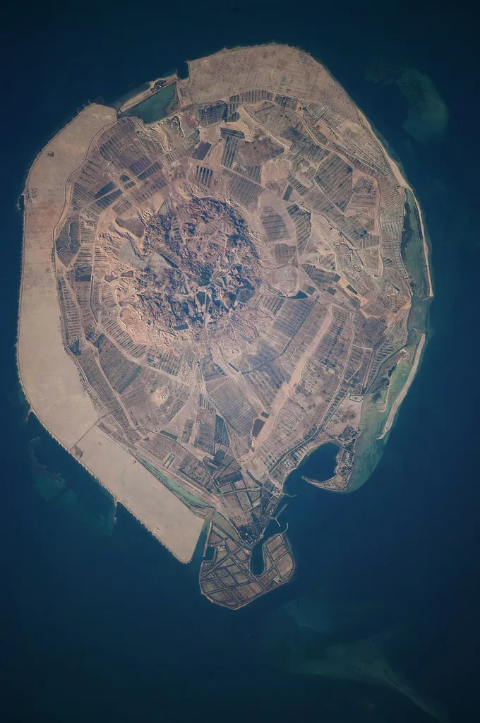 This astronaut photograph illustrates the varying character of surfaces on Sir Bani Yas. The central mountains of Jebel Wahid (image centre) mark the location of the Sir Bani Yas salt dome. The dome has breached the surface but exposed salt, primarily gypsum, is eroded, leaving a rugged, insoluble cap made of fragments of the overlaying sedimentary and volcanic rocks. Sand and silt derived from the Jebel Wahid and surrounding gravel-covered areas form beaches along the outer edge of the island. Tan graded areas along the western and north-eastern coast of the island (image bottom and image left) may be re-vegetated with additional plots or developed for other land uses.