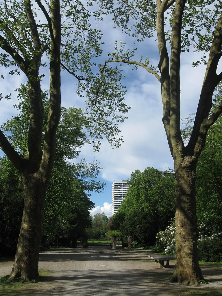 Blick von der Allee durch den Haupteingang des Gelsenkirchener Stadtgartens über Zepellinallee in Richtung Stadtzentrum auf den Stadtpark, im Hintergrund ist ein Teil des Hotelturms (Maritim-Hotel) zu sehen.