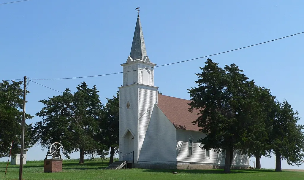 Dannevirke Danish Lutheran Church, at the northeast corner of county roads Dannevirke Road and Wausa, northwest of Elba in Howard County, Nebraska; seen from the southwest. The church was built in Gothic Revival style in 1906. With the community hall on the southwest corner of the intersection, it constitutes the Dannevirke Danish Lutheran Church and Community Hall, which property is listed in the National Register of Historic Places. The red cedar trees in the photo were planted in 1907. At lower left is the bell that formerly occupied the steeple; it was removed after the steeple was struck by lightning in 1994.